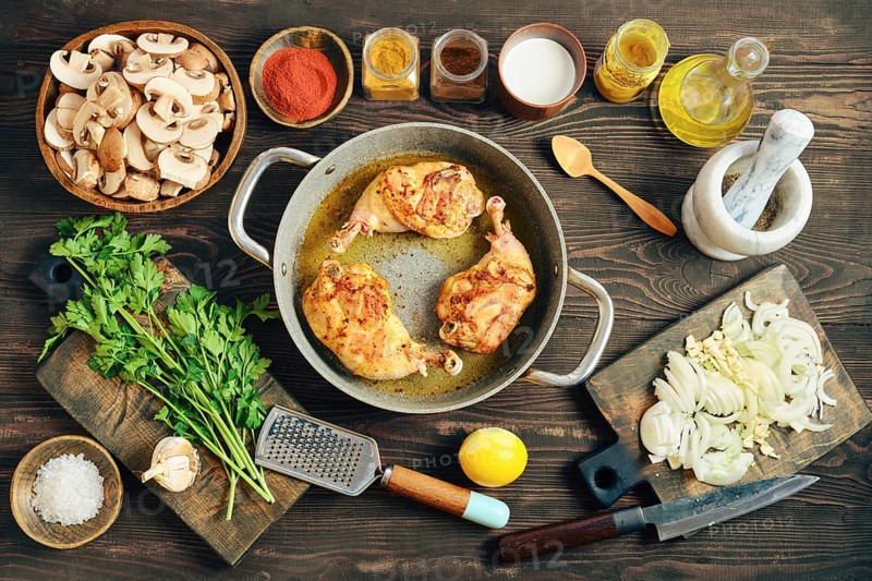 Roasted chicken legs in a pan on a wooden tabletop surrounded with mushrooms, onions, and various spices. Fresh herbs and a lemon are also on the table ready for use