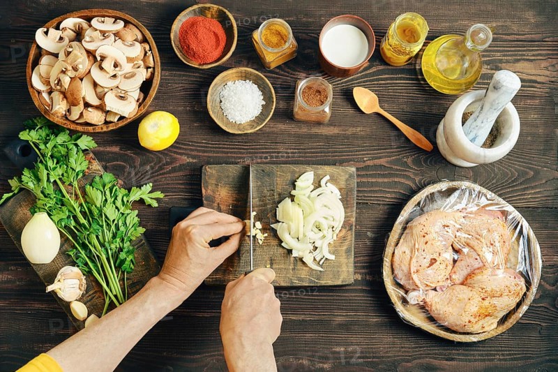 High angle view of hands chopping fresh garlic on a wooden cutting board. Chopped mushrooms, marinated chicken thighs, spice and herbs are located on the table