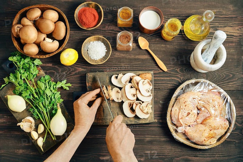 High angle view of person cutting mushroom for a meal. Lifestyle food preparation with wrapped chicken, onion, and herb on a dark rustic kitchen table