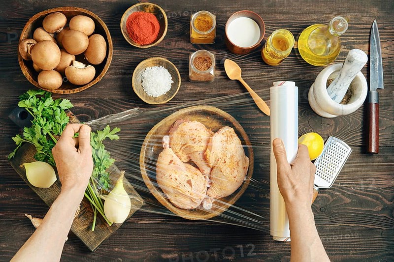 Hands wrapping marinated chicken with plastic stretch film for marinating. High angle view of rustic wooden tabletop with ingredients