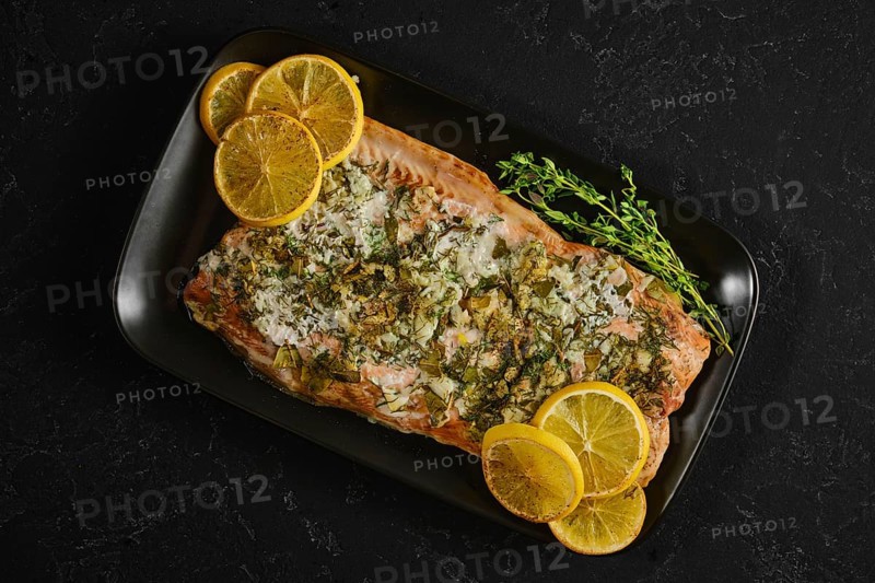 Roasted pink salmon is served on a black plate with slices of lemon and herbs. This dish is ready for tasting during dinner or a special occasion. The vibrant colors add appeal to the meal