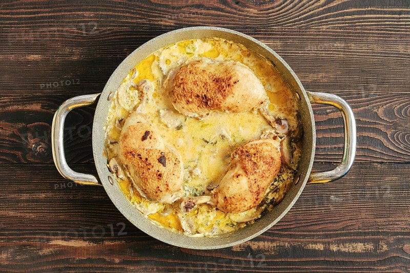 Tree pieces of chicken in a pot filled with creamy mustard mushroom sauce and vegetables. The pot sits on a wooden table, and the scene is bright with natural light from outside