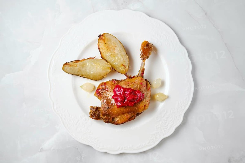 Succulent duck confit with crispy skin resting beside roasted pear caramelized in fat, garnished with a red sauce. The scene shows a meal ready to be enjoyed, highlighting traditional cooking methods