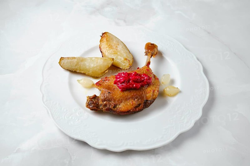 Duck confit served on a white plate with caramelized in duck fat pear wedges. A dollop of lingonberry sauce is on top of the duck. This dish is ready for someone to enjoy at a dining table