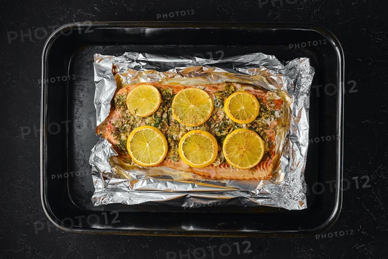 Pink salmon is placed on a baking sheet lined with foil. Lemon slices are arranged on top of the fish. The kitchen setting shows the preparation for a meal