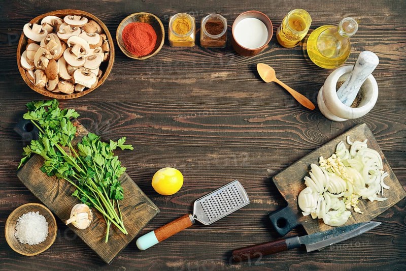 Fresh ingredients and tools for cooking are arranged on a wooden tabletop. Flat lay with chopped mushrooms, onions, herbs, spices, and kitchen tools for meal preparation