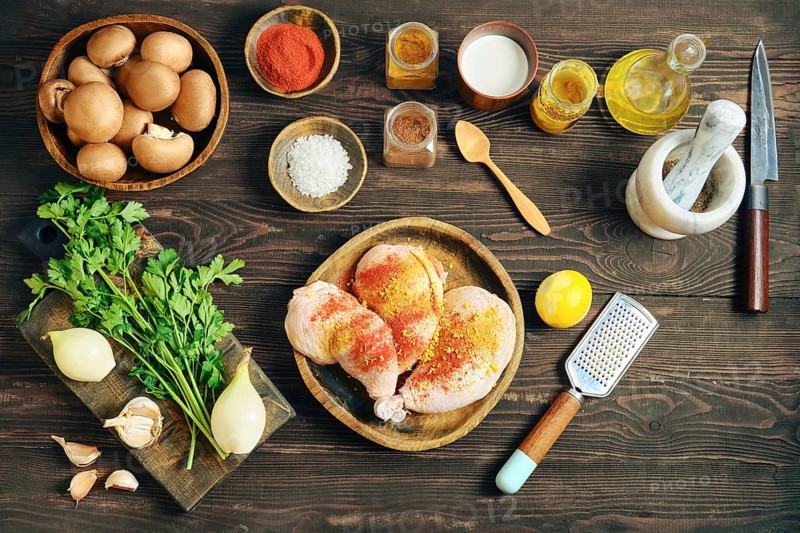 High angle view of raw chicken meat marinated with aromatic spice and lemon zest. Flat lay of home cooking process with mushroom, onion, and herb on a dark wooden table
