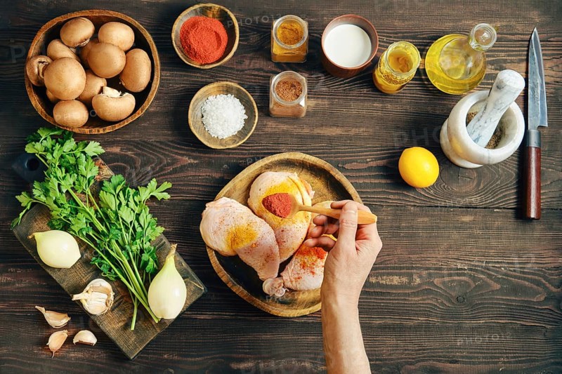Hand sprinkle smoked paprika over raw chicken thighs making dry marinade. Other raw ingredients for roasting are arranged nearby on wooden tabletop