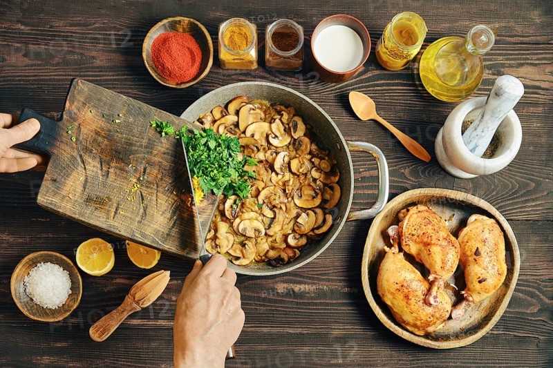 Unrecognizable person adds chopped herbs to a frying pan with fried sliced mushrooms. Nearby, bowls of spices, lemon, and roasted chicken are visible. Process of making creamy mushroom sauce