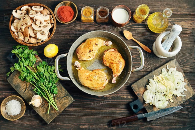 Top view of chicken poultry browning in oil. Culinary process of making dinner with sliced mushroom, onion, and garlic on a dark rustic background