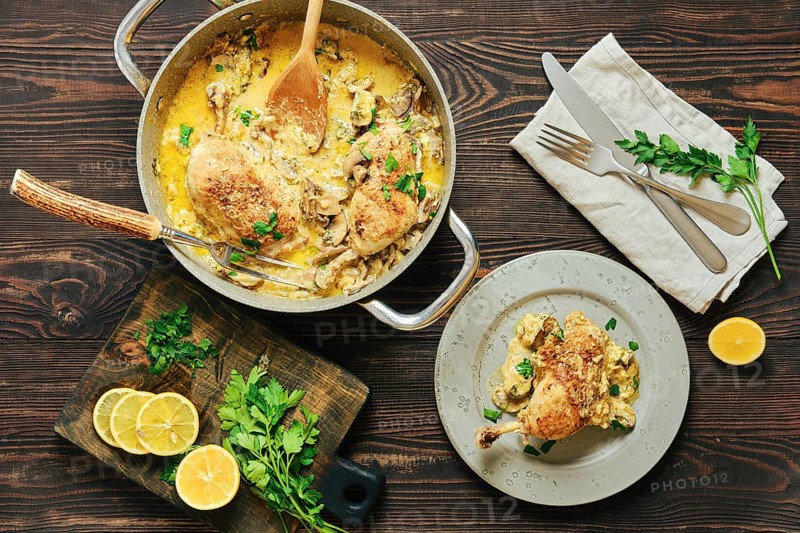 Top view of home-cooked creamy chicken legs in mushroom sauce served with parsley and lemon on rustic wooden table