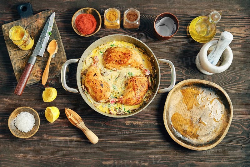 Three pieces of chicken cooked in a pot with a mix of spices and creamy mustard sauce. The pot is on a table surrounded with spices and other ingredients like lemon, salt, and oil