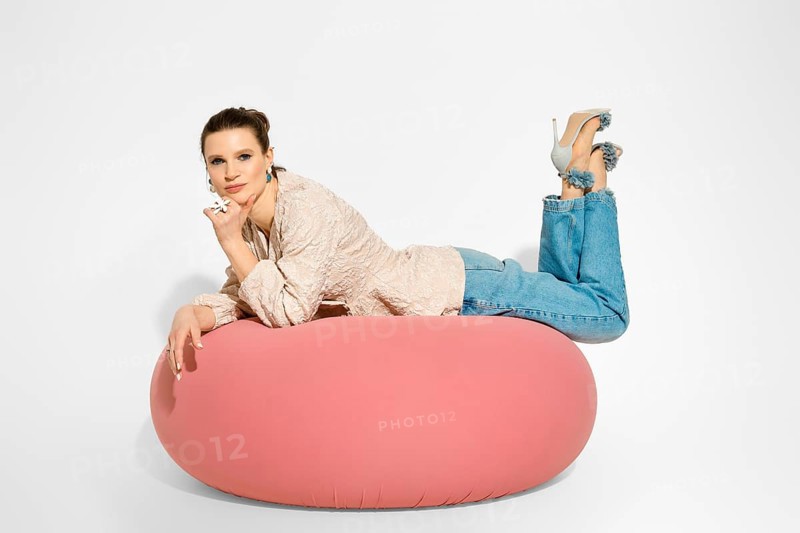 A model lies on a pink cushion with her elbows resting on hand. She wears a light top and denim jeans. Her feet, in heels are crossed