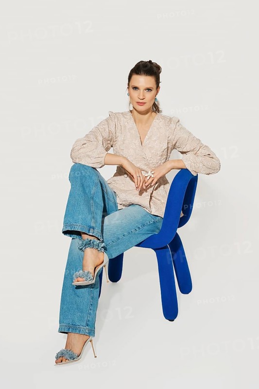 A woman sits on a blue chair with one leg crossed over the other. She wears a light top and denim jeans. The setting is a studio with a simple background, focusing on her outfit and pose