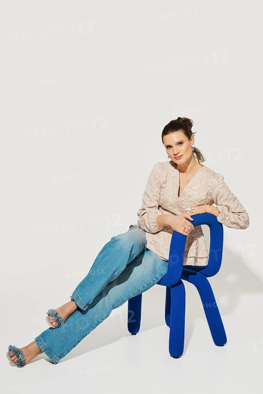 A woman is sitting on a blue chair with her legs extended. She wears a light top and denim jeans. The background is plain and the setting is bright. It is clear that it is daytime