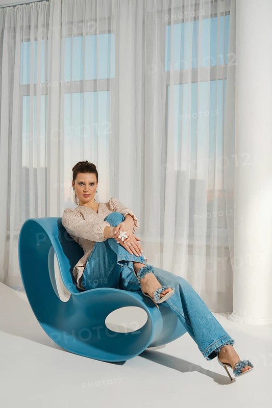 A woman is sitting on a blue chair. She is wearing casual clothes and is smiling. The room has a lot of natural light coming in through the windows. She looks relaxed and confident
