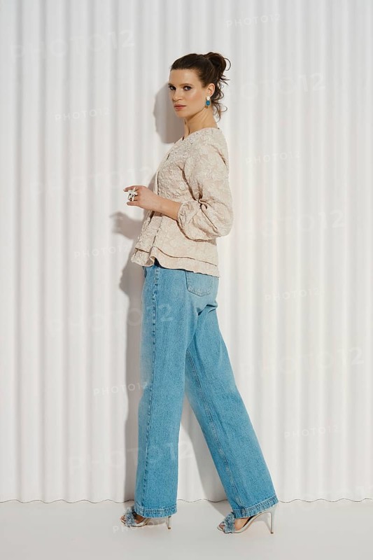 A model stands sideways with her left foot forward, wearing a casual outfit consisting of a light top and blue jeans. She is posed in front of a white striped backdrop
