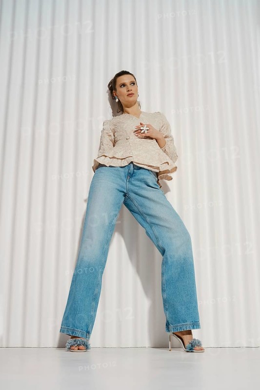 A woman stands confidently in a relaxed pose wearing wide-leg jeans and a light blouse. She is in a modern indoor space with a textured white wall. Her styling is simple, showcasing her outfit