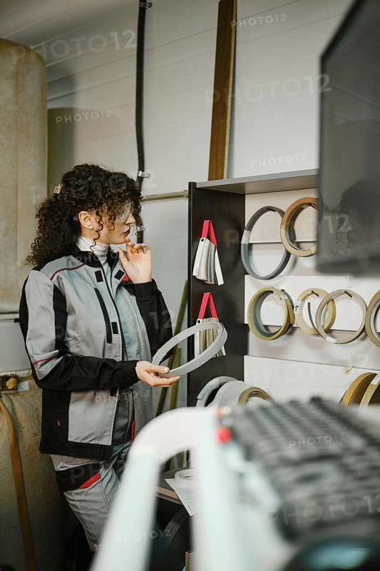 In a workshop, a woman looks closely at finishing edges displayed on a shelf. She stands by equipment and checks the variety of sizes. Her work clothing and safety glasses show a focus on her task