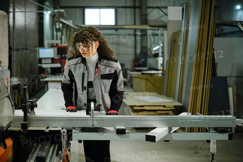 A person stands at a workbench in a workshop, preparing to cut wood. The space is filled with tools and raw materials. The worker wears safety gloves and a gray work suit