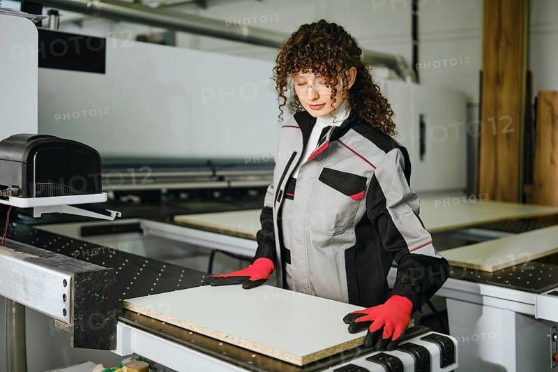 A person stands at a machine in a wood processing facility, wearing work gloves and a jacket. She is working with wooden panel ready for cutting, surrounded by machinery and equipment