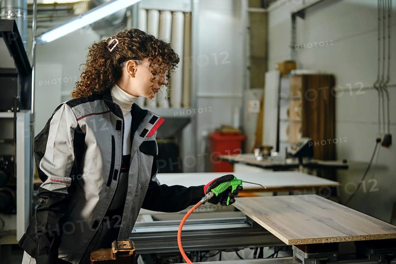 Female carpenter use a spray gun while blowing away dust from a wooden panel placed on a milling machine