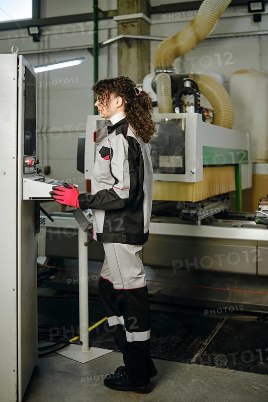 Woman stands at a control panel inside a factory, wearing protective clothing and gloves. The worker is focused on operating machinery while tools and equipment are working nearby
