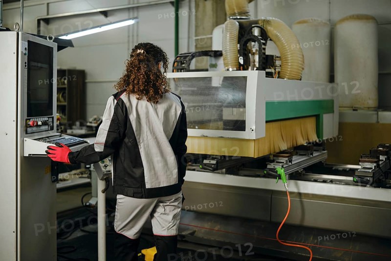 A woman uses a milling machine in a workshop. She stands with her back to the viewer and watches the milling machine. The environment is well-lit with various tools and equipment in the background