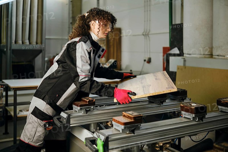 A woman operates a machine in a carpentry workshop. She is laying a wooden panel on a milling machine. She wears protective gear and handles the material with care