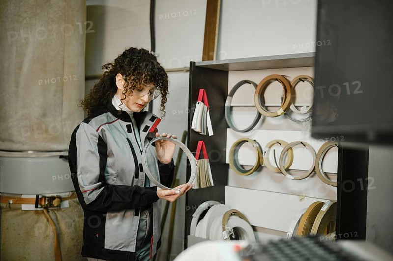 A woman stands in a workshop looking closely at a finishing edge. She is surrounded by various types of edge tapes displayed on a shelf