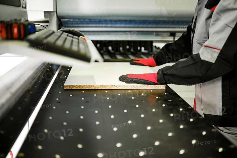 A person in gloves operates a machine that cuts a piece of material in a workshop. The workspace has several tools and equipment. Focus is on precision and safety during the cutting process