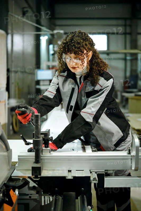 A worker stands at a machine in a factory, using tools to operate equipment. The person wears safety gear such as goggles and gloves