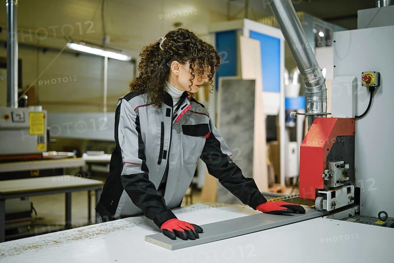 A woman with curly hair works at a woodworking machine in a workshop. She wears gloves and a jacket. The space has wood pieces and tools, and bright lights are on