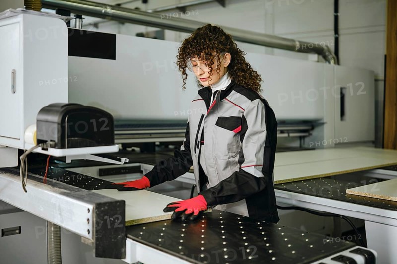 A woman stands at a CNC machine in a factory. She puts a wooden sheet in a sawing machine. She is focused on her task while wearing gloves and safety glasses