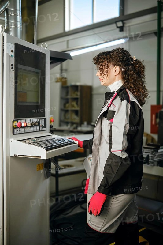 Woman stands at a control panel in a manufacturing facility. She wears a work uniform with gloves. The machine is active, showing data on the screen