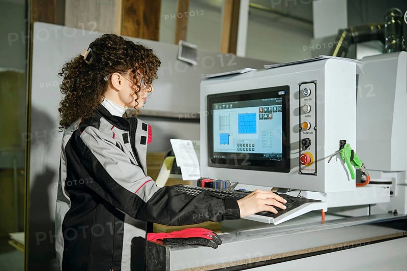 A woman using a computer to control cutting machine at furniture factory. She wears work clothing and focuses on the screen