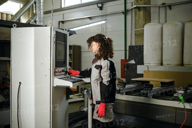 A person operates a milling machine in a carpentry workshop filled with tools and equipment. The worker wears gloves and stands by the control panel, focused on the task