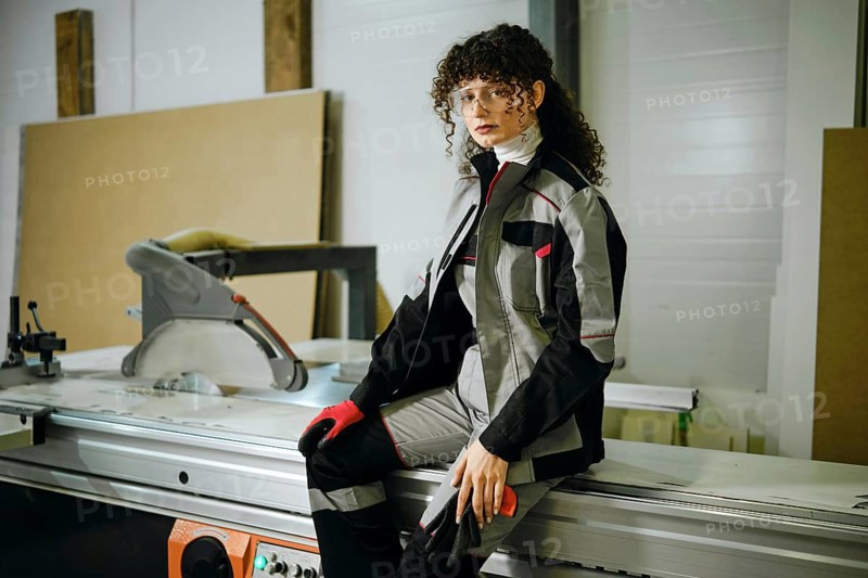 A woman sits on a machine in a workshop. She is wearing a work outfit and gloves. The workshop has tools and equipment around her. Natural light comes through the windows