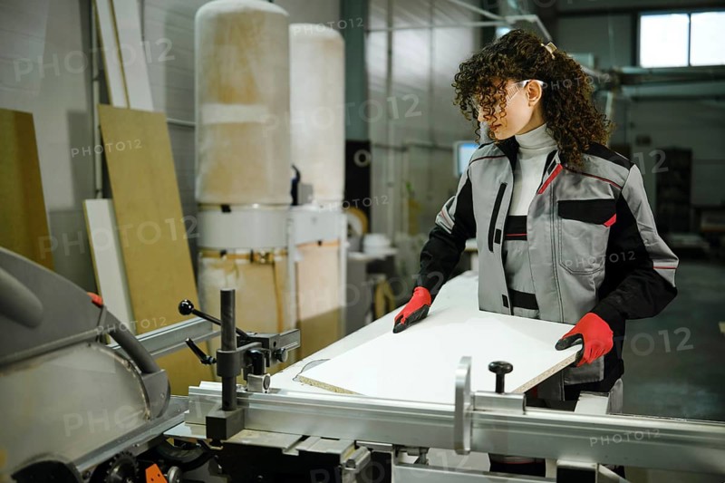 Female worker stands in a workshop focused on cutting a wood board with a table saw. The person wears safety gear like gloves and goggles. The space has equipment and materials all around
