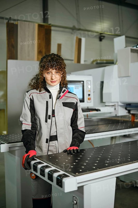 A woman stands in a workshop next to a wood shaping machine. She wears a work suit and gloves while looking at the camera. The space is filled with equipment and materials