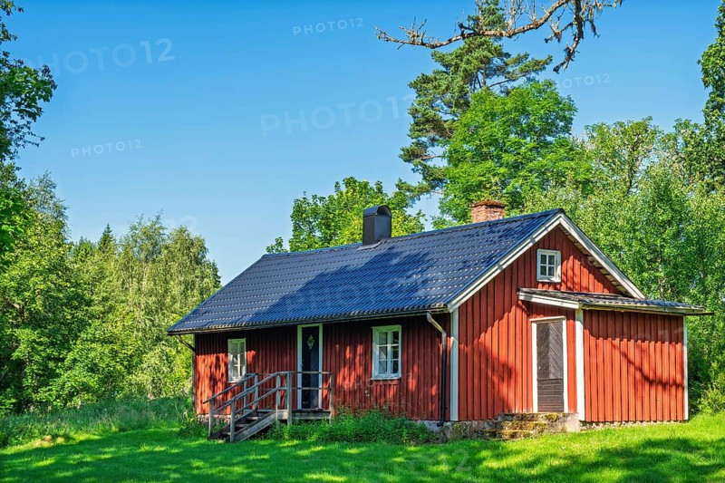 Red wooden country house with dark roof on green lawn surrounded by trees and forest under clear blue sky a sunny summer day, Sweden