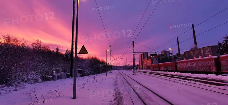 Sunrise at a snow-covered train station on Rennsteig, railroad tracks in purple sky with factory in the background, Frankenwald nature park Park