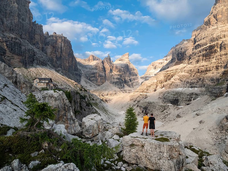 Two hikers in front of impressive mountain peaks of the Brenta Mountains, evening at Refugio Francis Fox Tuckett, Brenta, Parco Naturale Brenta-Adamello, Trentino, Italy