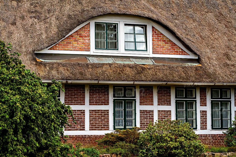 Traditional thatched house with brick walls and several windows, surrounded by green vegetation, The façade of a historic thatched house near Stade