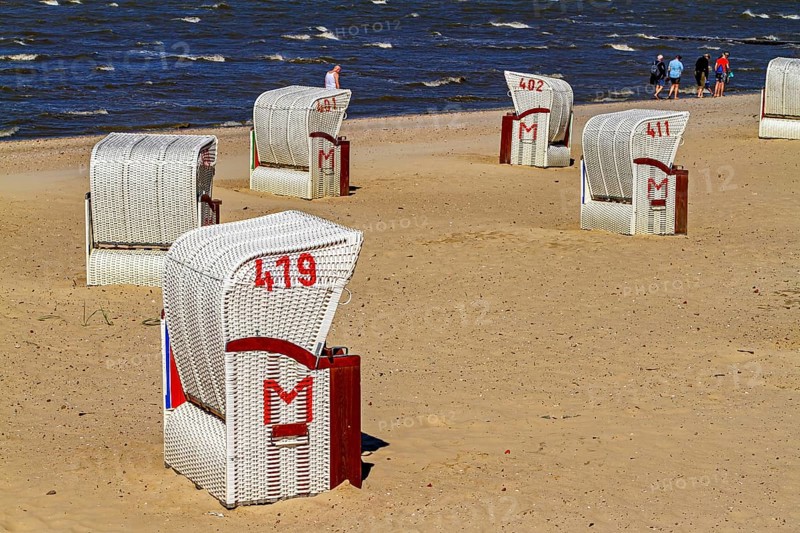 Several white beach chairs with red accents stand on the beach with walkers in the background, beach chairs on the beach in Cuxhaven
