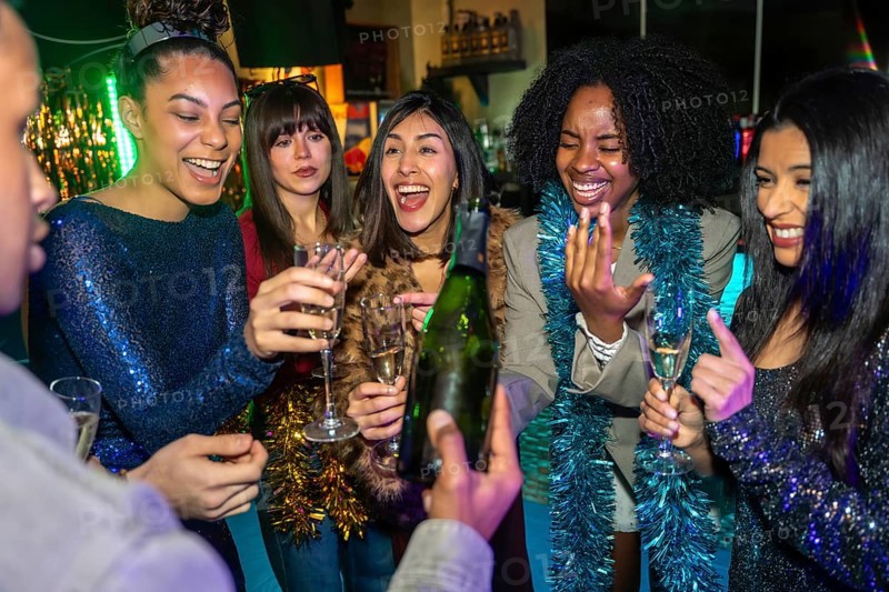Group of excited diverse young adult friends smiling and laughing while celebrating a special event, enjoying a bottle of champagne and sparkling drinks at a lively party