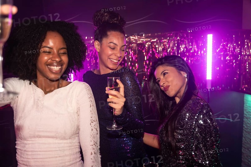 Three diverse women friends enjoying a night out, toasting with champagne and smiling while celebrating in a vibrant nightclub under purple neon lights with tinsel decorations