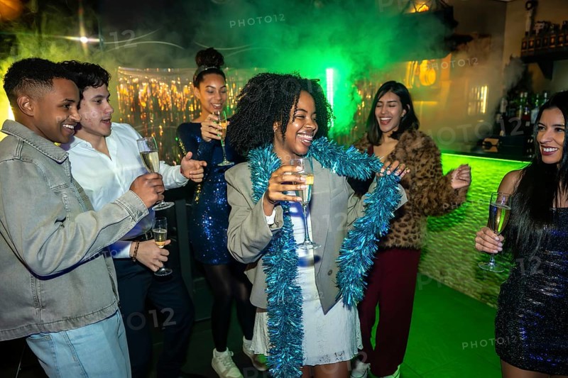 Group of diverse young adults having fun, dancing, and laughing while holding champagne flutes at a vibrant night club or bar, celebrating a special occasion with festive lights and smoke