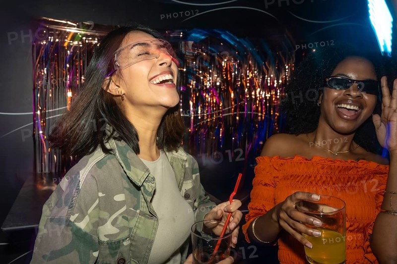Two diverse young women enjoying a night out, laughing and dancing with drinks in hand amidst colorful club lights and festive decor, celebrating friendship and fun