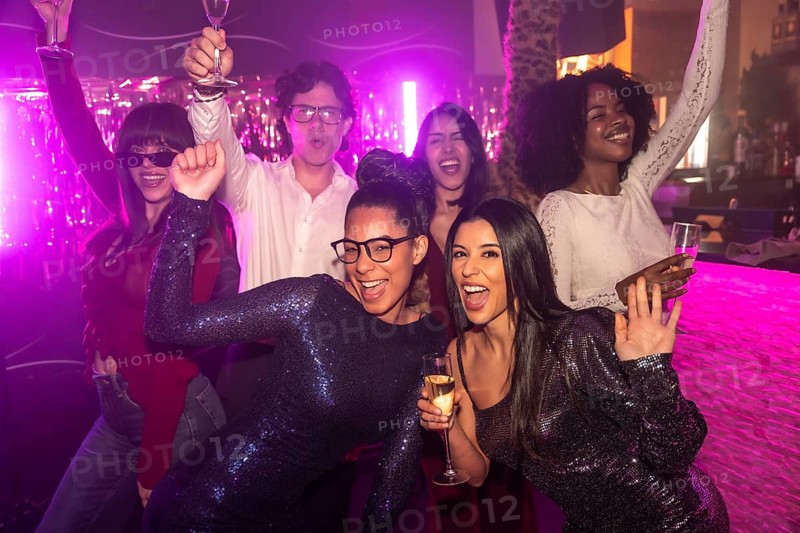 Diverse group of young adult friends dancing and celebrating together at a vibrant nightclub, holding drinks and smiling, illuminated by pink and purple neon lights